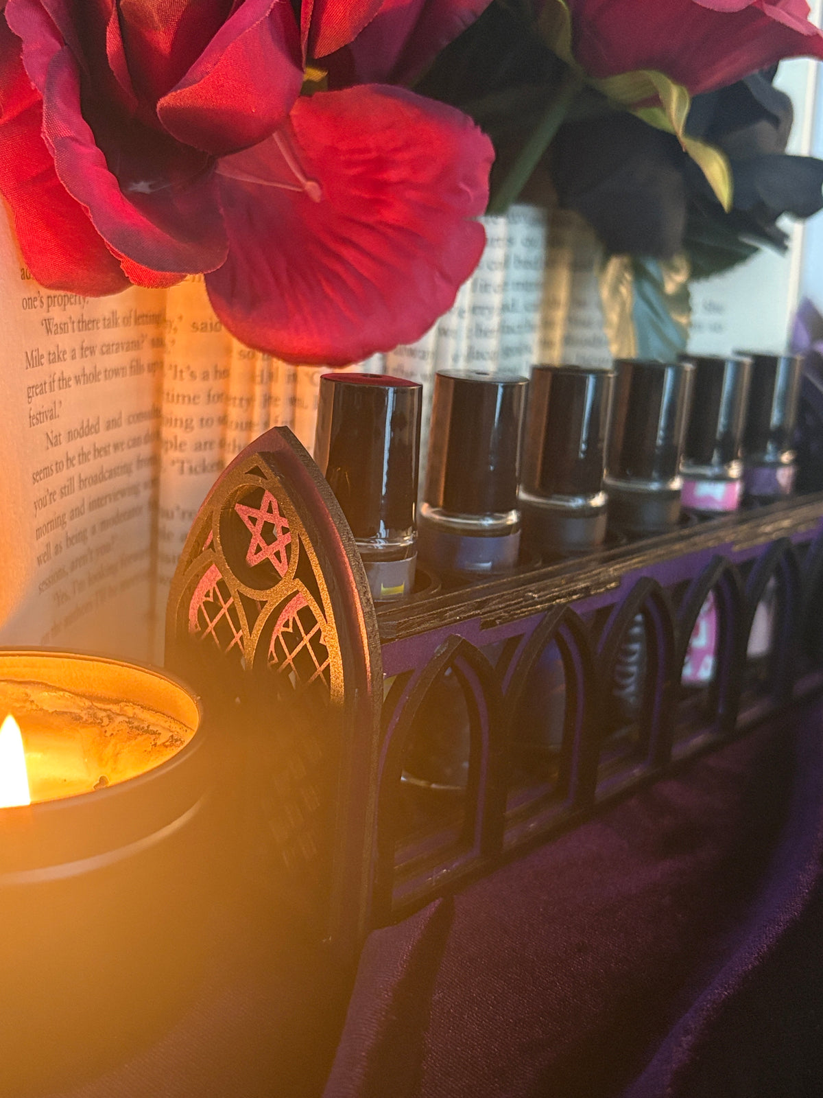 A close-up of a black pentacle holder filled with small black bottles, styled with red flowers, a burning candle, and a book in the background.